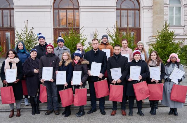 Zdjęcie przedstawia laureatów Stypendium pod Choinkę stojących na tle choinek oraz Gmachu Głównego PW